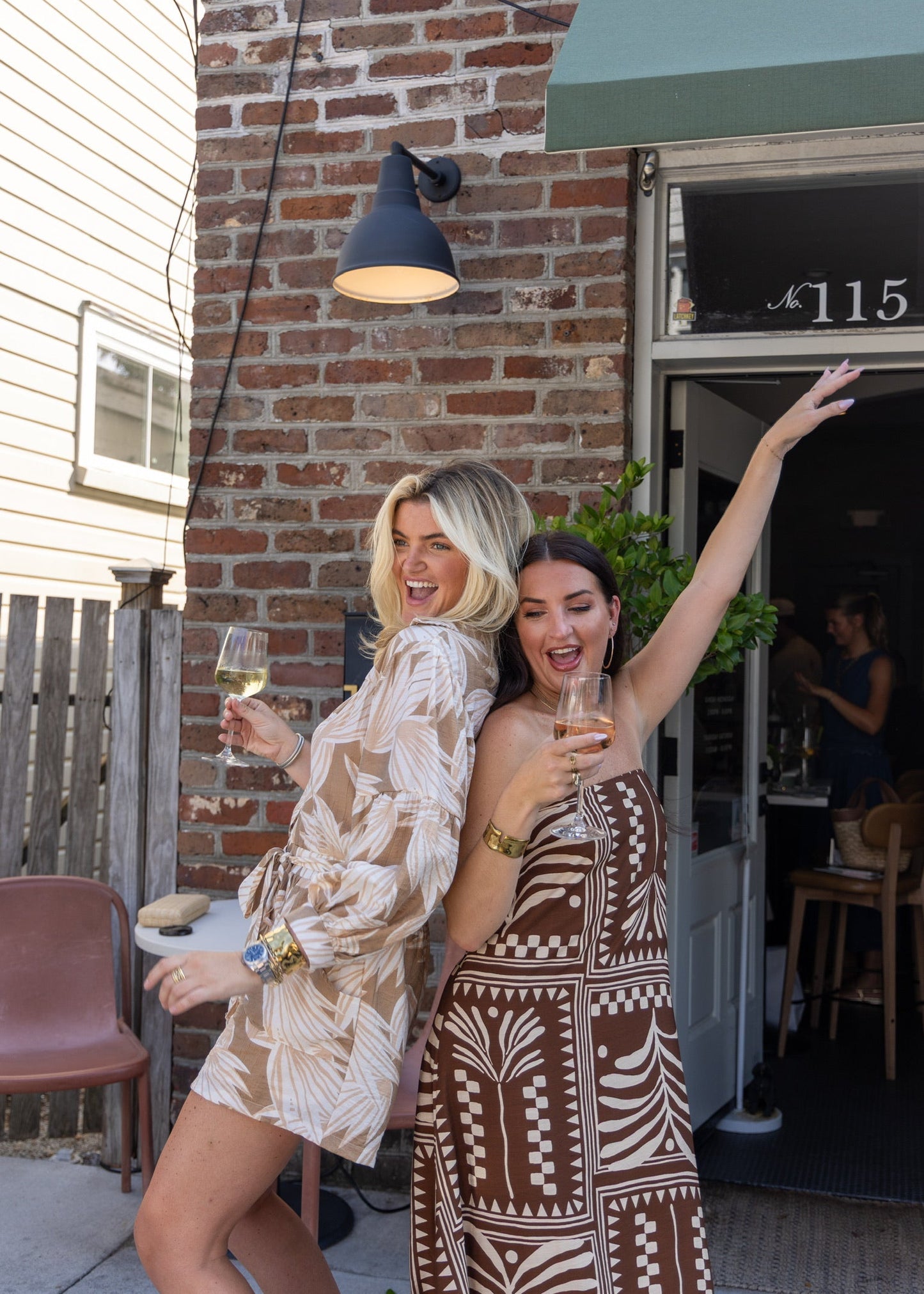 two women with wine glasses celebrating their new outfits. wearing a rompter in tan and a long brown dress with a cream geometric print