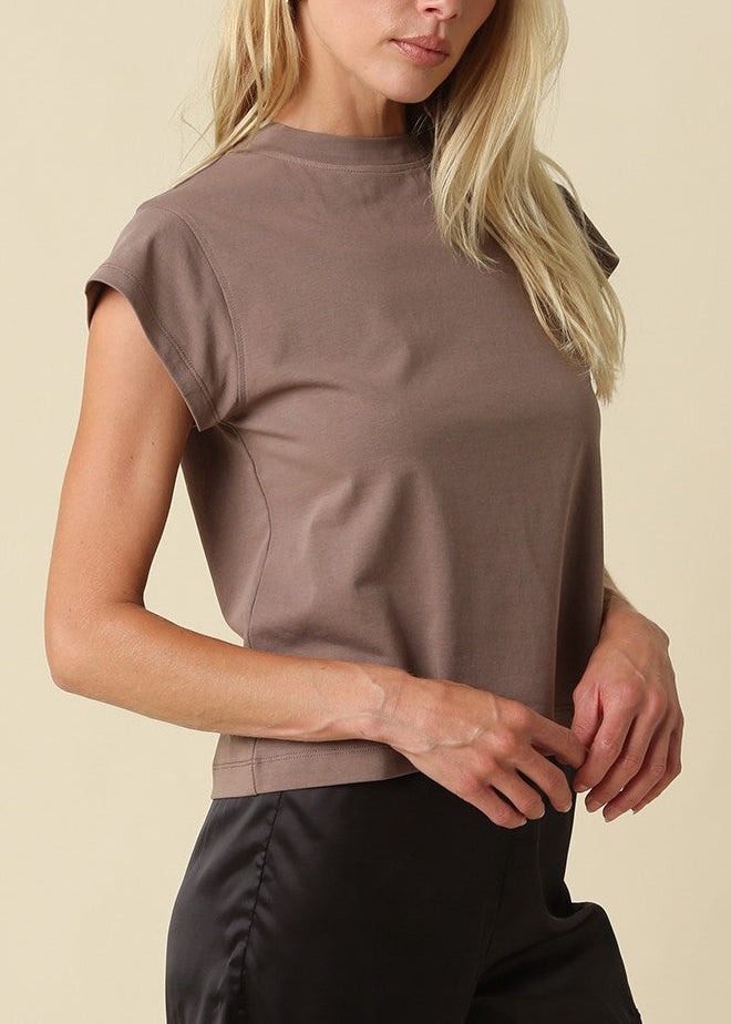 A blonde woman wearing a taupe-brown short-sleeve mock neck t-shirt and black pants, posing against a solid beige background.