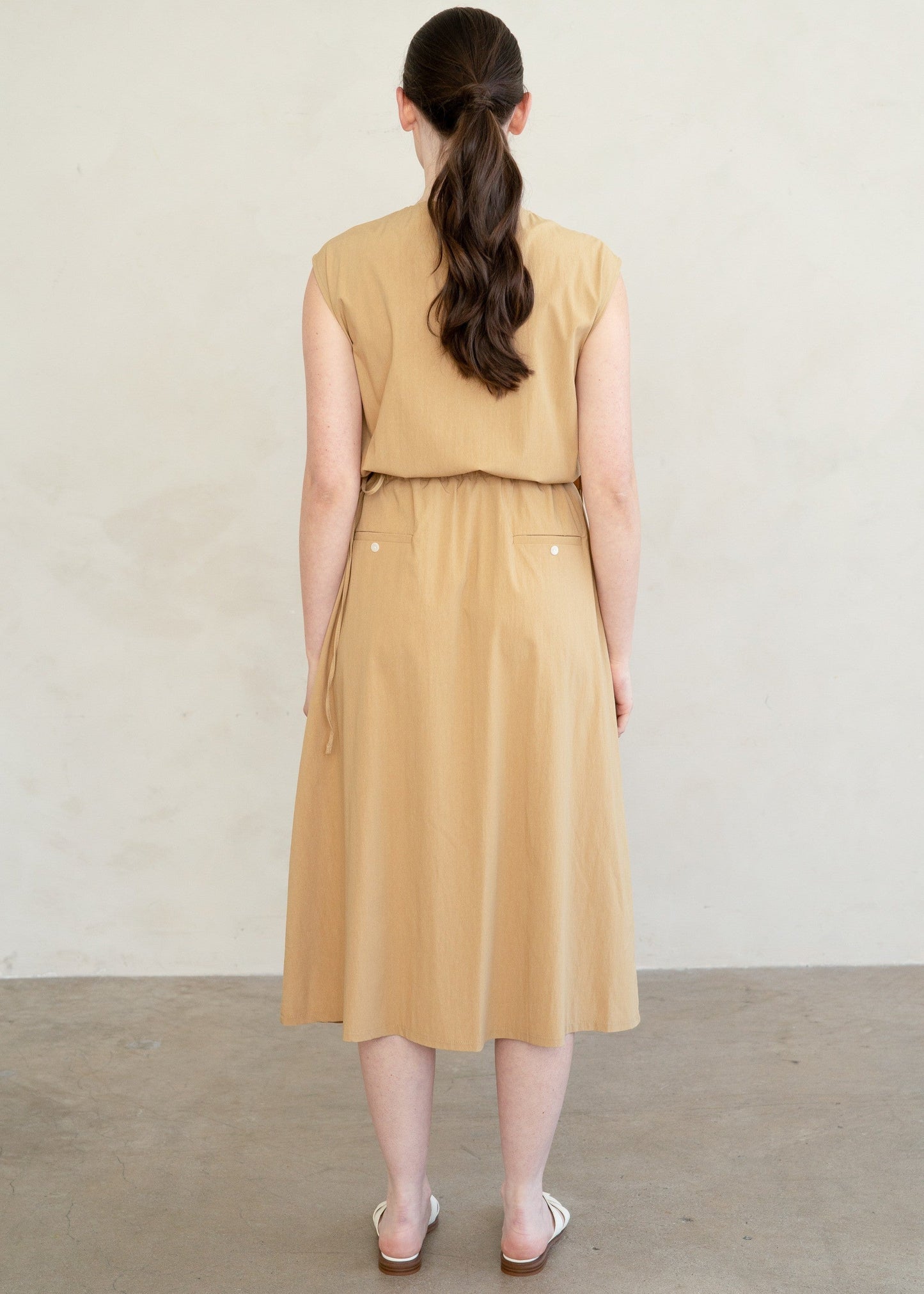 A woman viewed from behind wearing a sleeveless mustard yellow dress with a tie waist and a midi-length skirt, standing against a neutral off-white wall.
