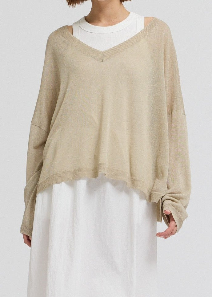 A model wearing a beige knit long-sleeve sweater layered over a white V-neck top, paired with a white maxi skirt and black sandals, against a plain light gray studio background.