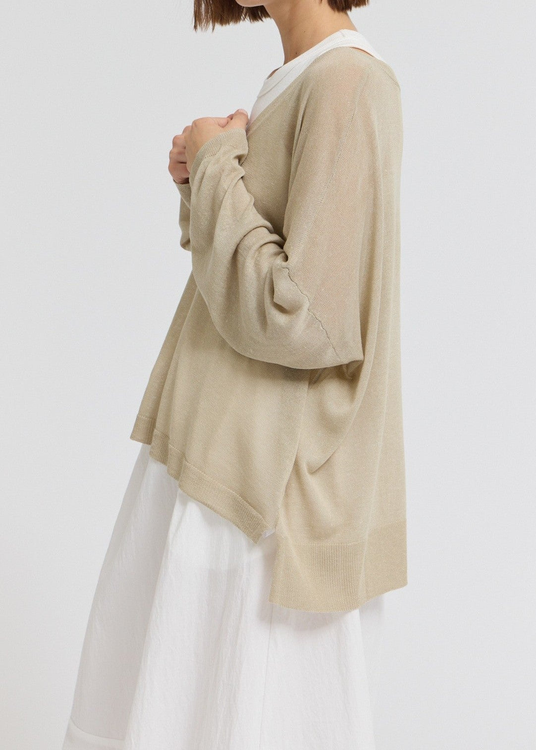 A model wearing a light beige, long-sleeved knit cardigan over a white top and white skirt, shown from a side profile against a plain white studio background.