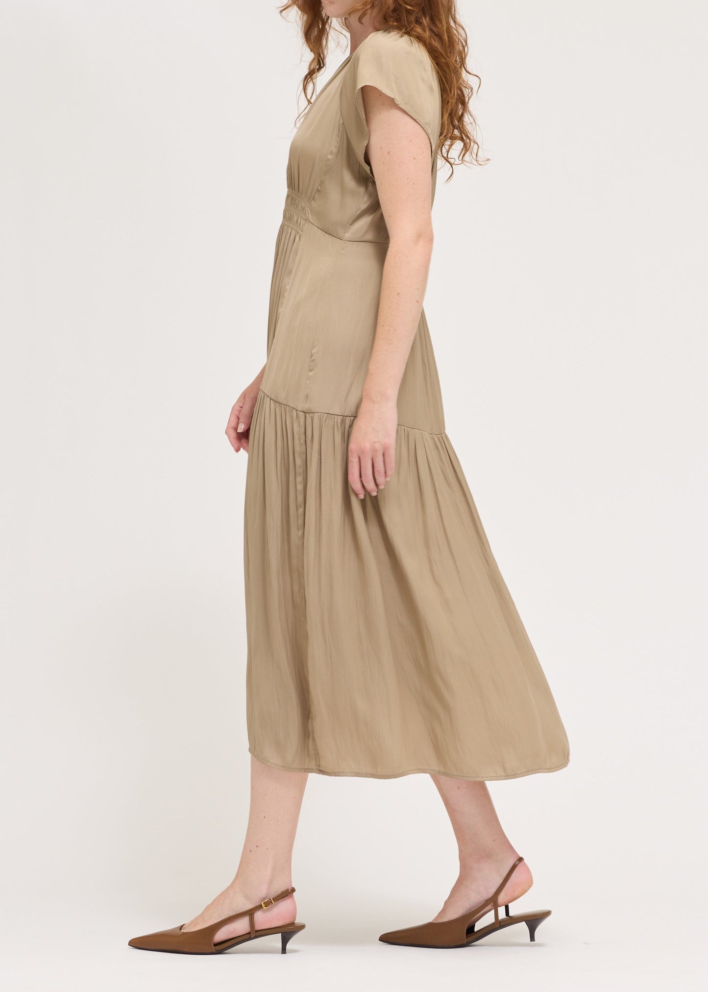 A woman with long wavy red hair is shown in profile wearing a beige midi dress with short sleeves and brown slingback kitten heels, set against a plain light gray studio background.