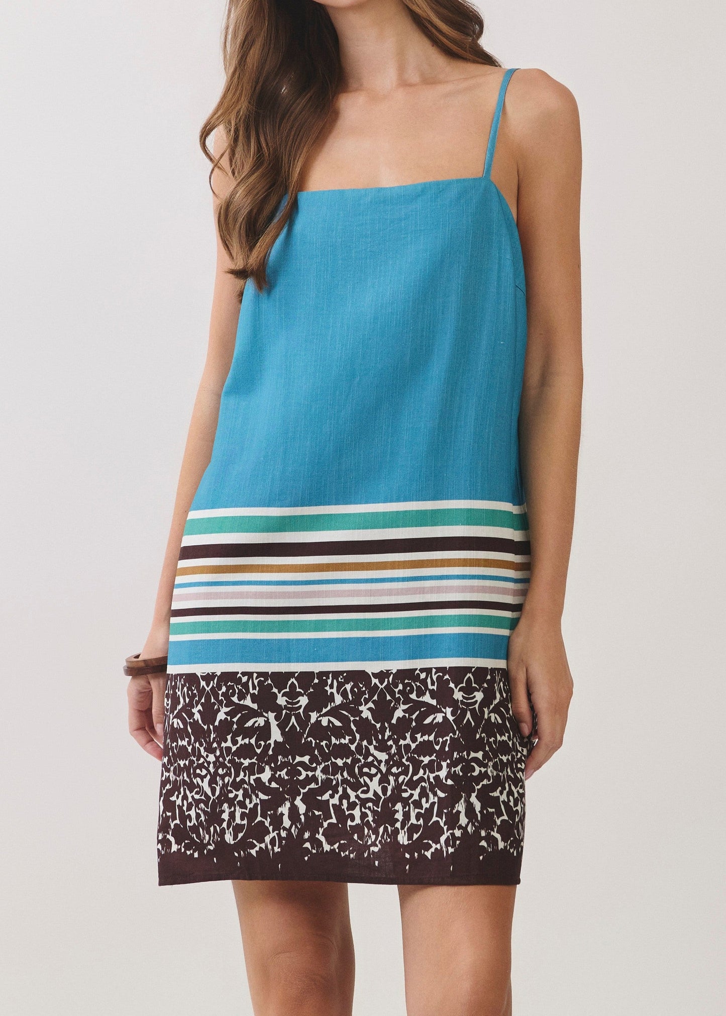 A woman modeling a sleeveless spaghetti-strap dress with a solid turquoise blue upper section, horizontal multicolor stripes across the waist, and a brown and white speckled lower skirt, set against a plain light gray studio background.