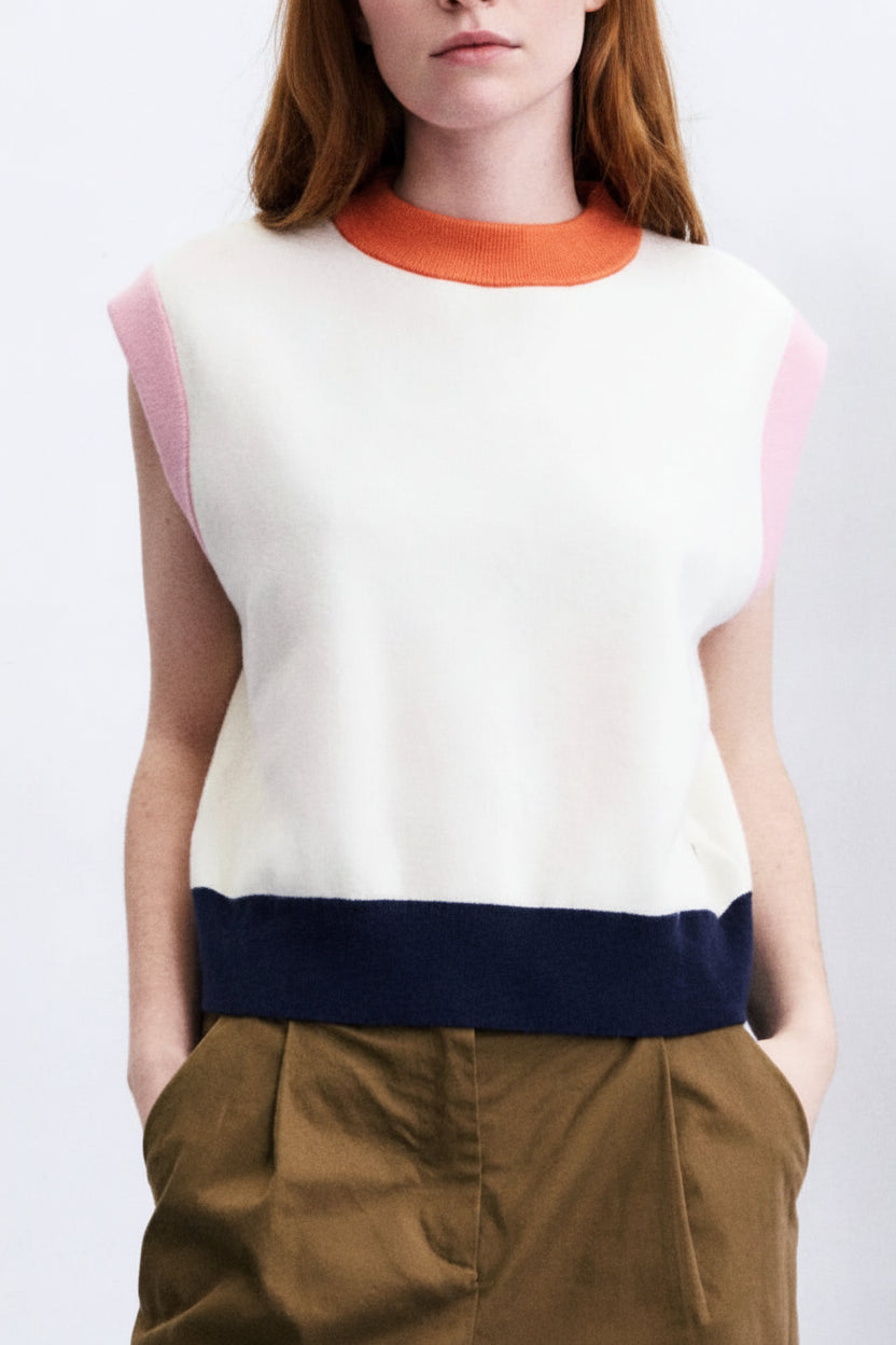 A woman stands in a studio wearing a sleeveless knit top with a cream body, orange collar, and pink shoulder panels, paired with dark navy and brown wide-leg pants. A tall rotating photo display stand is beside her against a white wall and blue floor.