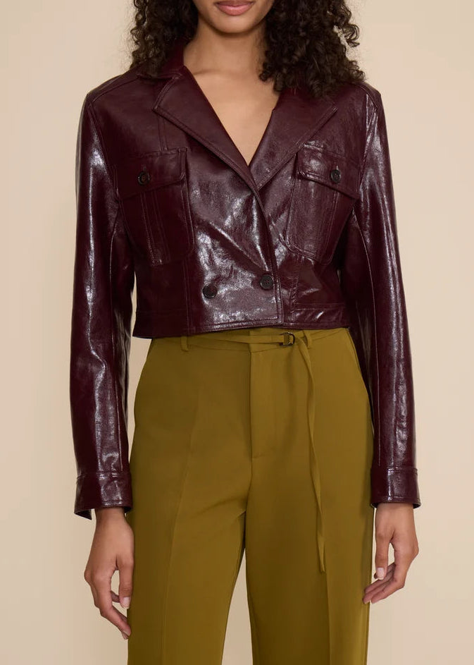 A woman modeling a cropped, glossy burgundy faux-leather jacket with a wrap-style front, paired with olive green trousers, against a solid beige background.