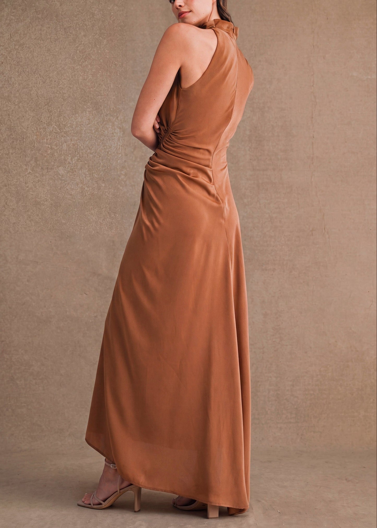 A woman modeling a floor-length, one-shoulder draped dress in a warm camel brown color, photographed against a textured neutral studio backdrop.