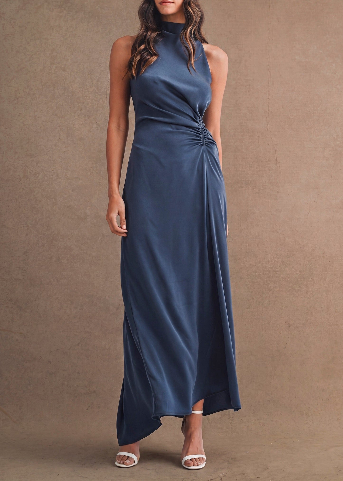 A woman modeling a sleeveless navy blue maxi dress with a high mock neck, side twist detail, and draped skirt, standing against a textured brown studio backdrop.