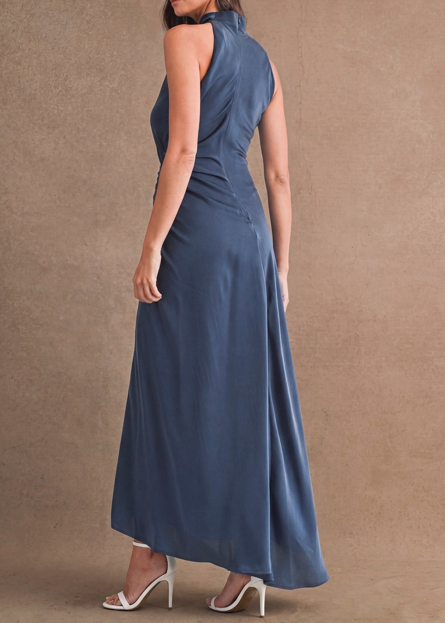 A woman modeling a sleeveless, high-neck, floor-length blue dress from the back, standing against a textured brown studio backdrop.