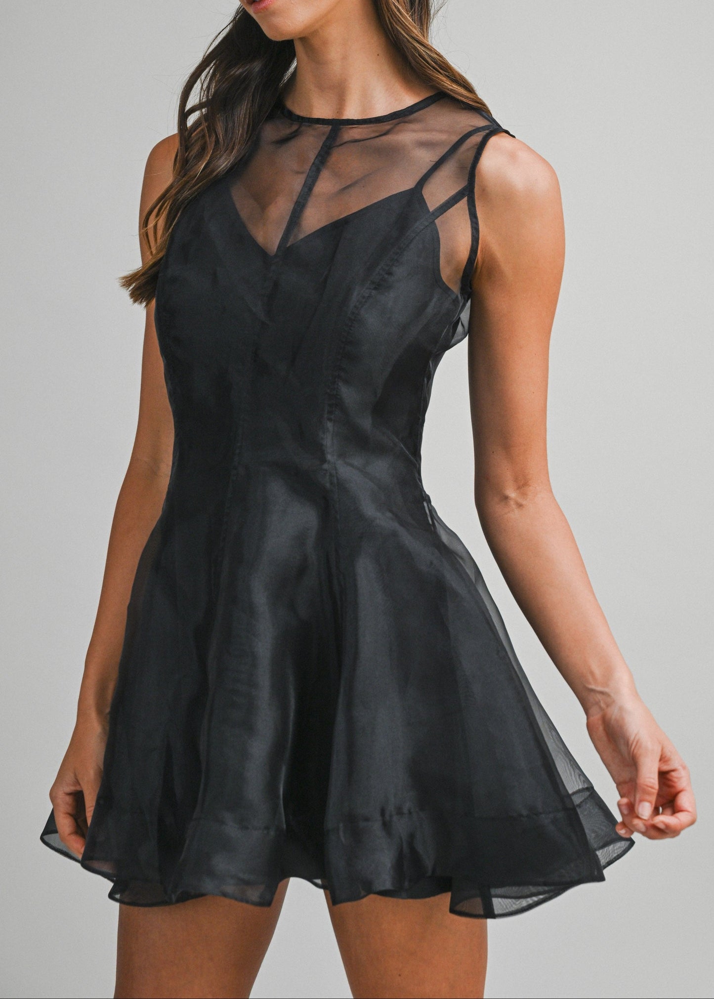 A woman modeling a black sleeveless mini dress with sheer mesh panels and thin shoulder straps, photographed against a plain light gray studio background.