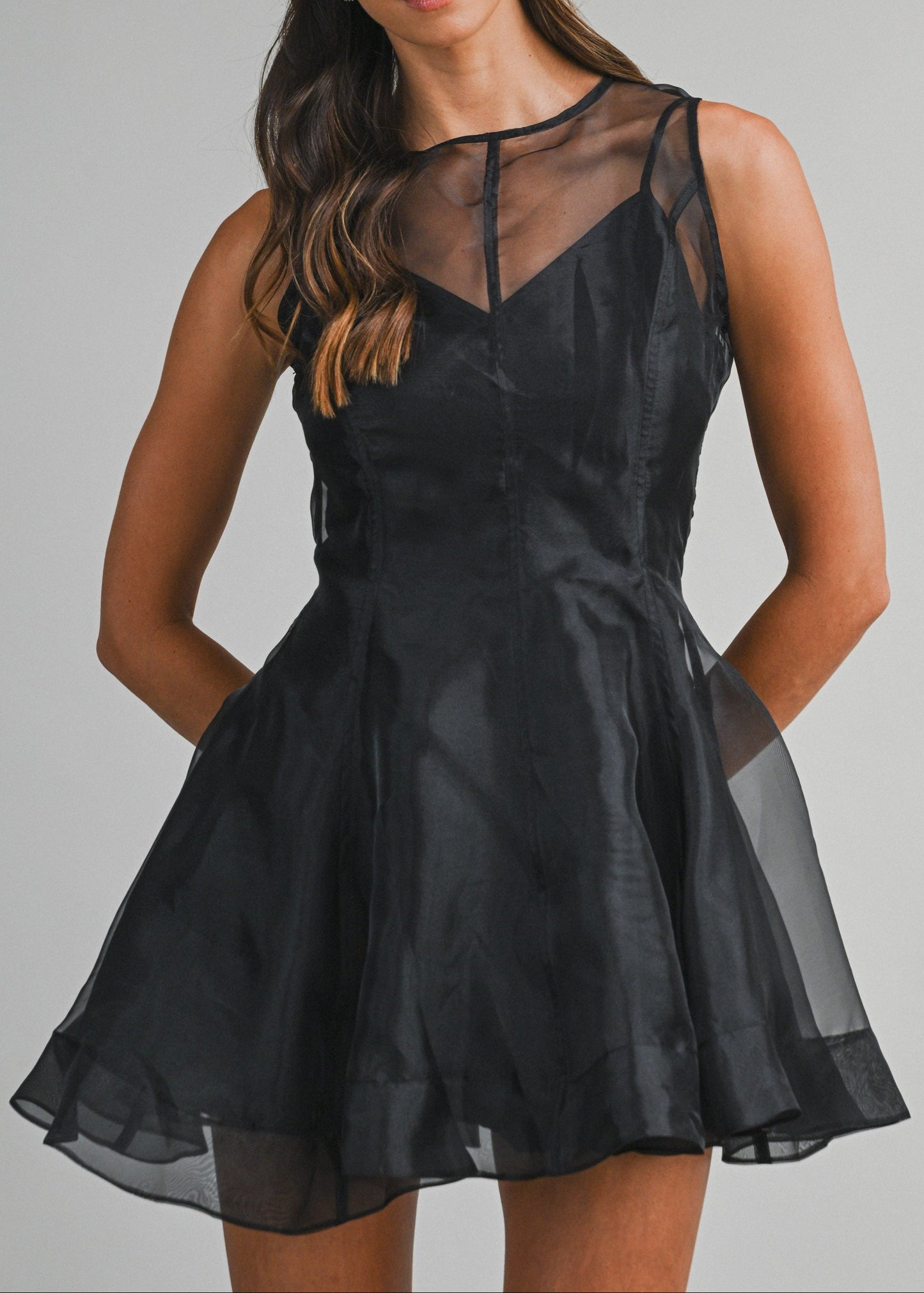 A woman modeling a sleeveless black dress with sheer mesh panels and a flared skirt, photographed against a plain light gray studio background.