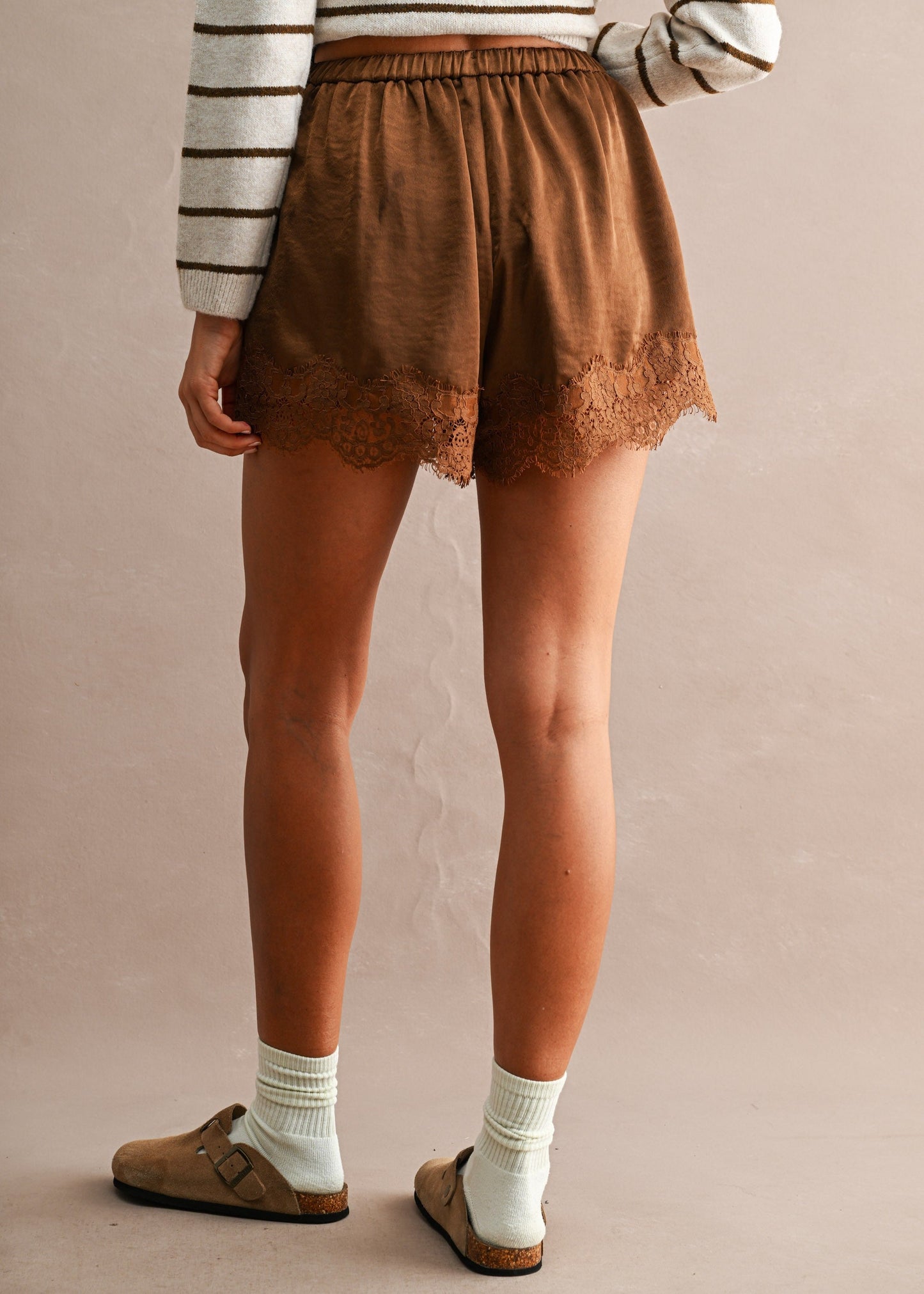 A person shown from the back wearing brown lace-trimmed shorts, a striped long-sleeve top, white socks, and brown clogs against a plain beige studio background.