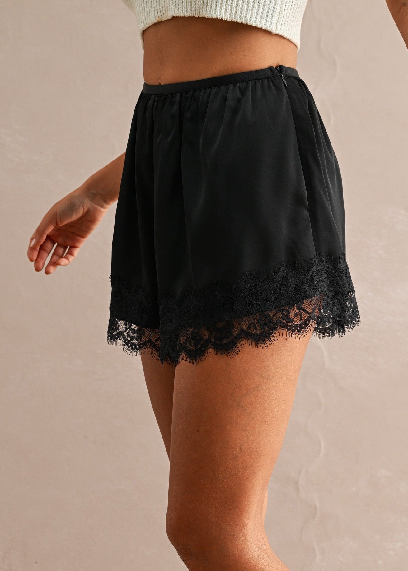 A model wearing a pair of black shorts with a lace hem, paired with black leather flats, shown from the waist down against a textured beige studio backdrop.