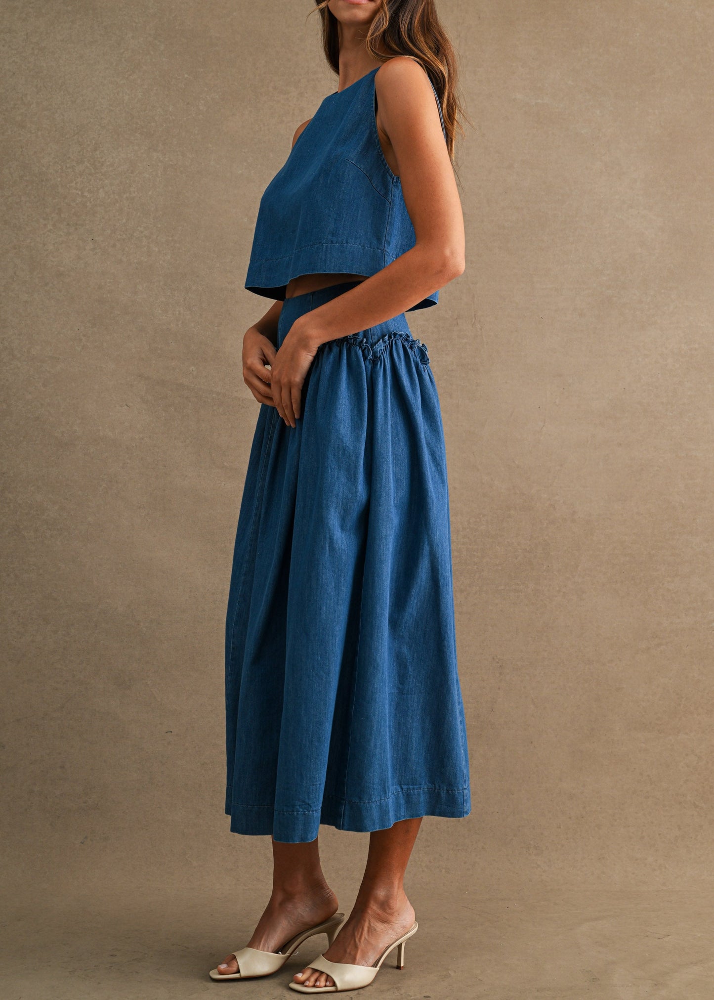 A woman modeling a two-piece blue outfit consisting of a sleeveless crop top and a high-waisted, wide-leg midi skirt, paired with white heeled sandals, set against a textured beige studio backdrop.