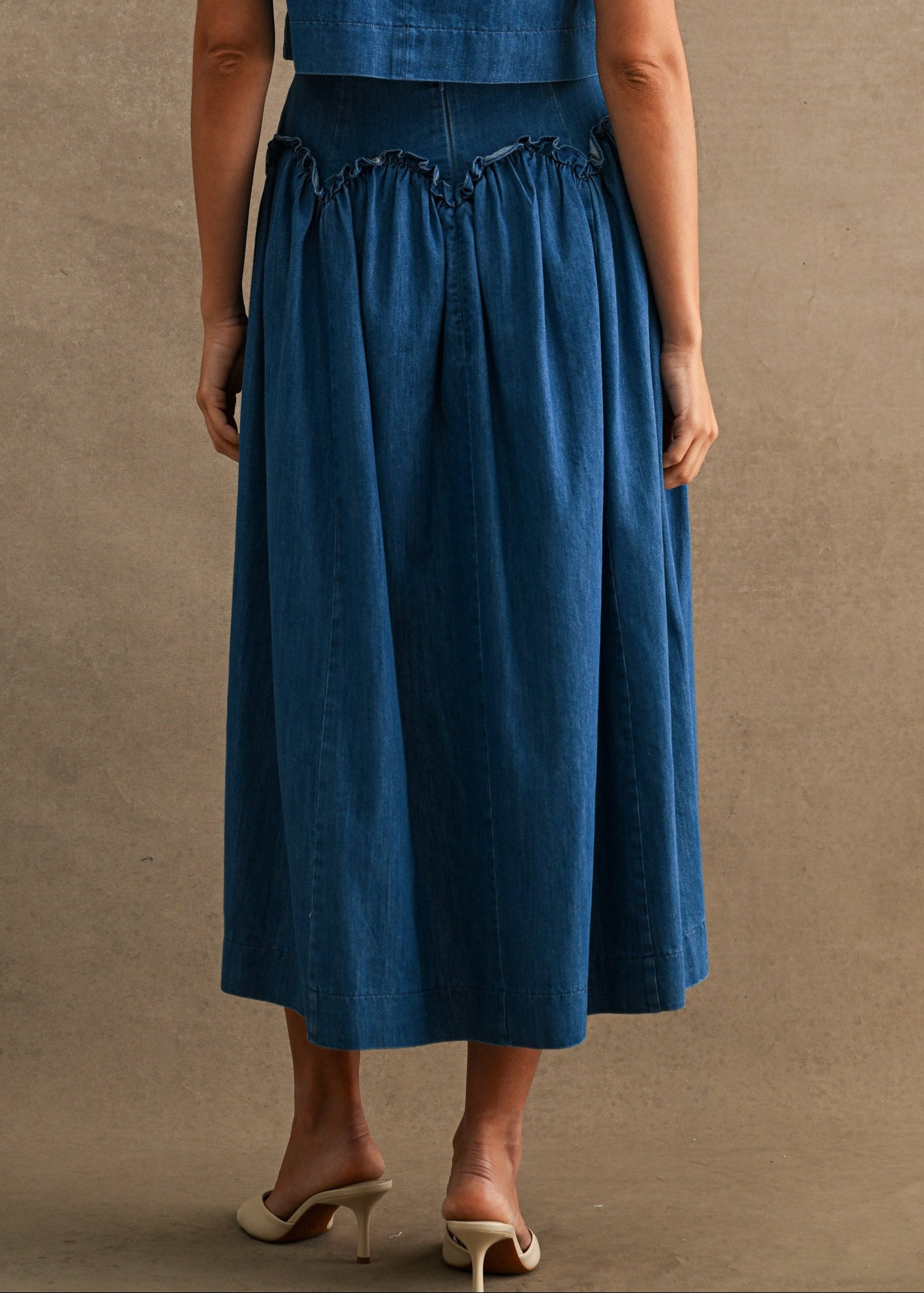 A woman viewed from the back wearing a sleeveless blue midi dress with a fitted bodice and gathered skirt, standing against a plain warm beige studio backdrop.