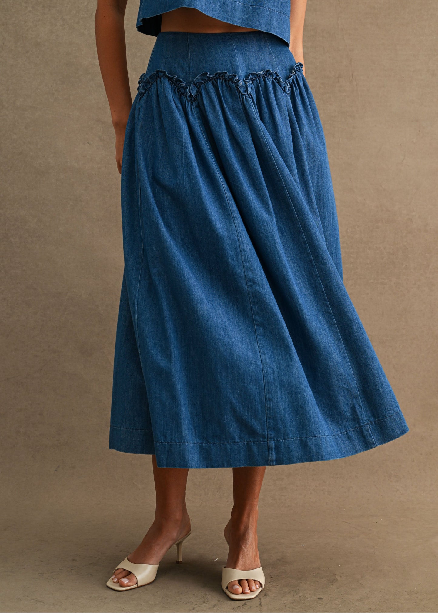 A model wearing a blue denim midi skirt with a ruffled waistband and a matching blue denim cropped top, styled with white heeled sandals against a textured beige background.