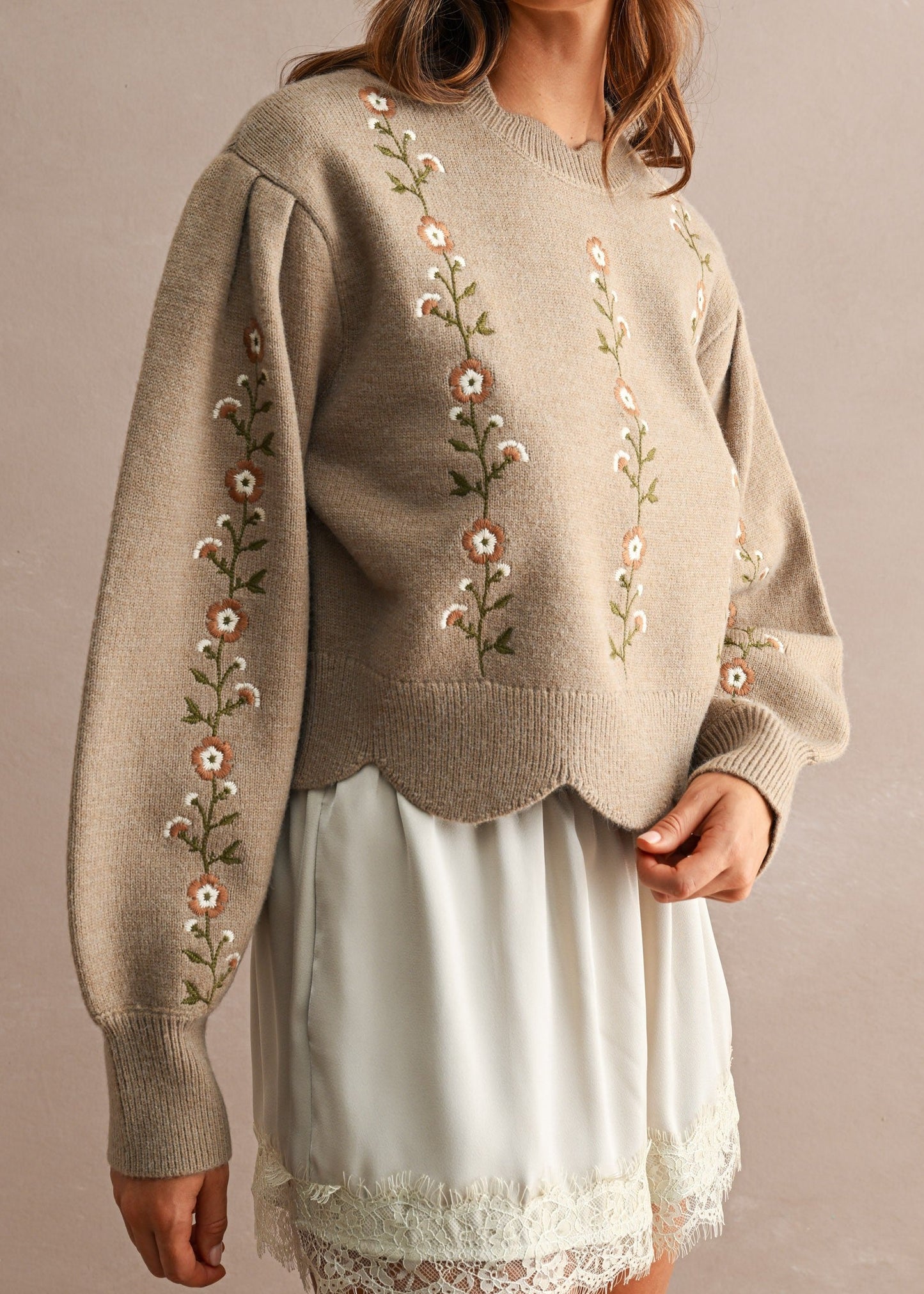 A woman wearing a beige knit sweater with floral embroidery on the front and sleeves, paired with a white skirt, standing against a neutral beige background.
