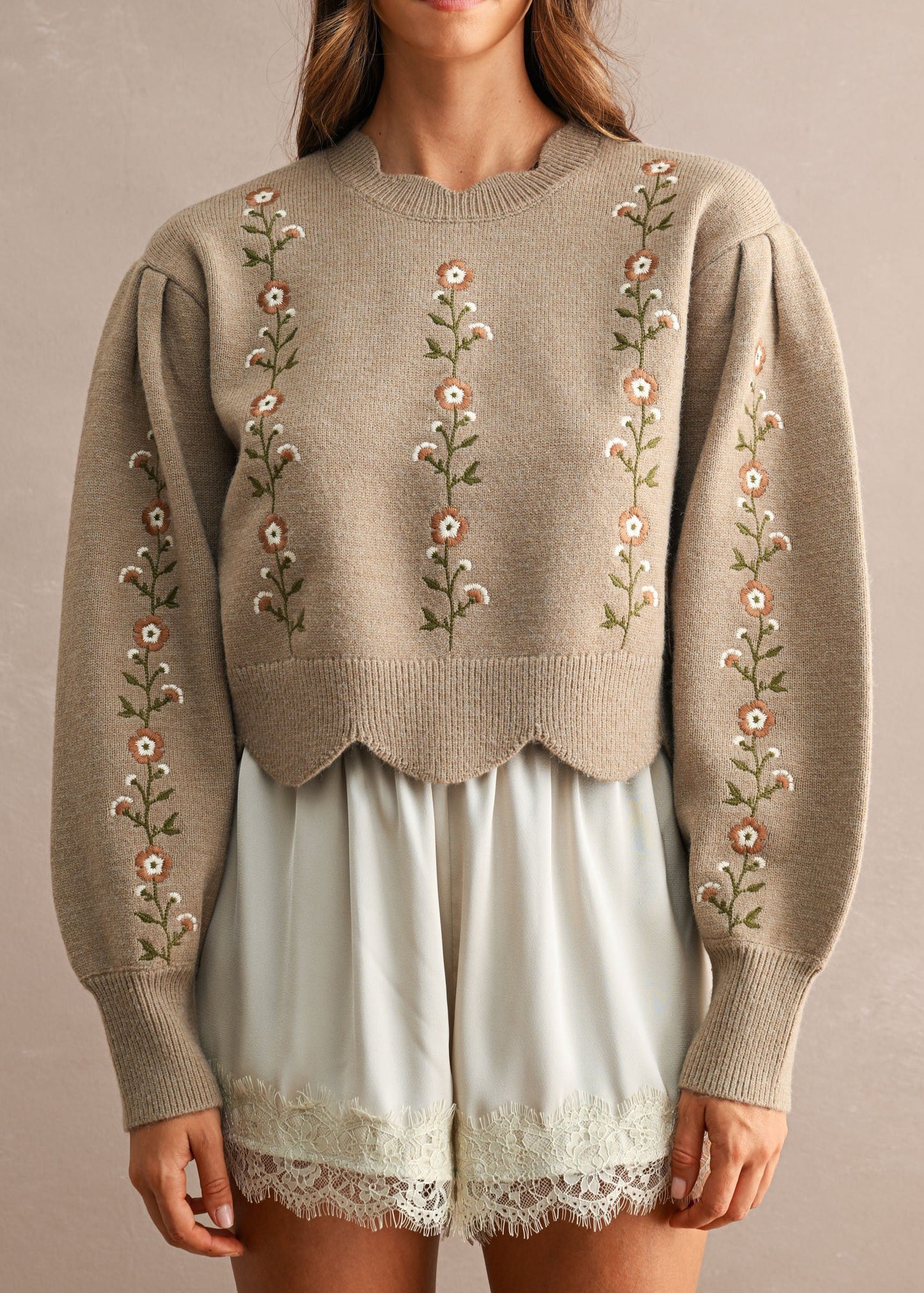 A woman wearing a taupe knit sweater with vertical floral embroidery down the front and along the sleeves, paired with light sage green shorts, against a neutral beige background.