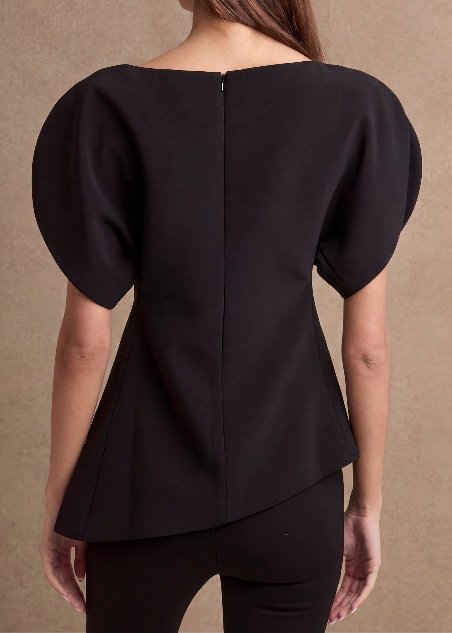 A woman viewed from the back wearing a black peplum-style top with short puff sleeves and a high-low hem, against a warm beige textured backdrop.