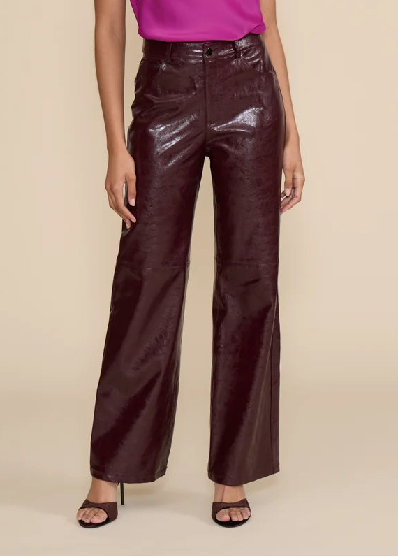 A model wearing high-waisted, wide-leg pants in a glossy, deep burgundy faux-leather material, paired with a pink top and black heeled sandals, against a solid beige background.