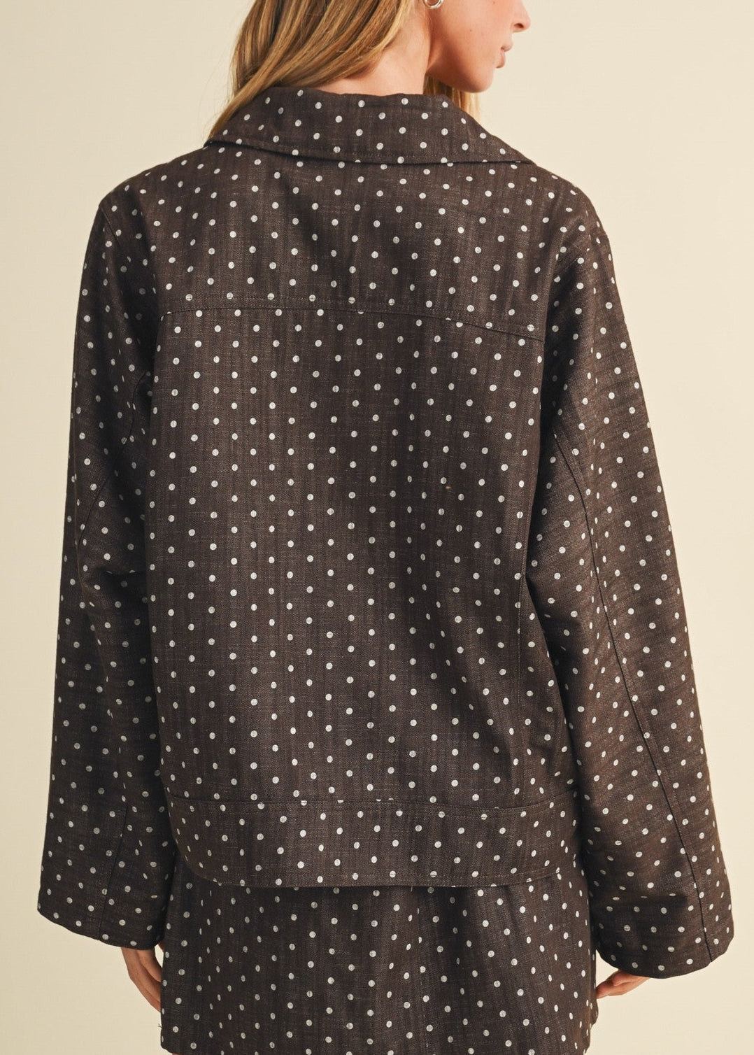 A woman shown from the back wearing a dark brown long-sleeve blazer with a white polka dot pattern, set against a plain beige studio background.