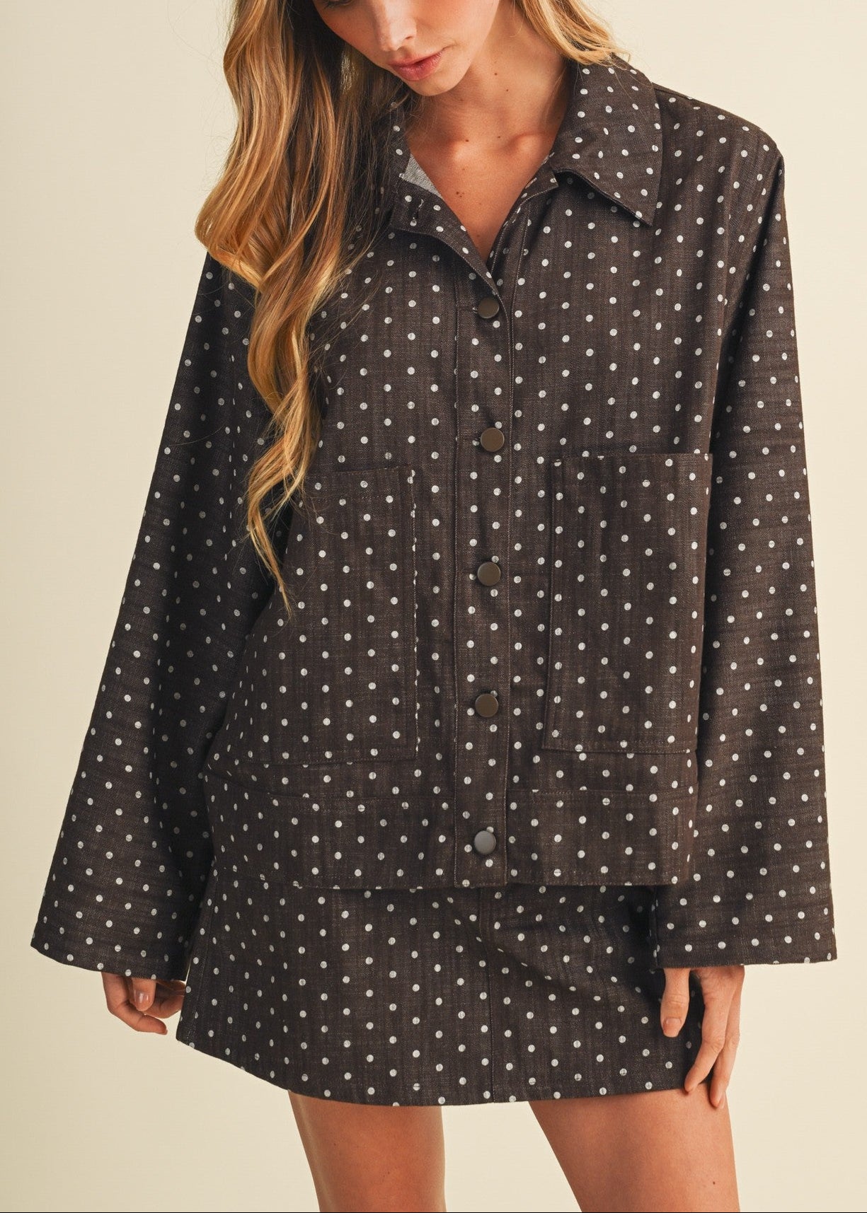 A woman wearing a dark brown long-sleeve button-up shirt and matching shorts set, both decorated with a small white polka dot pattern, set against a plain beige background.