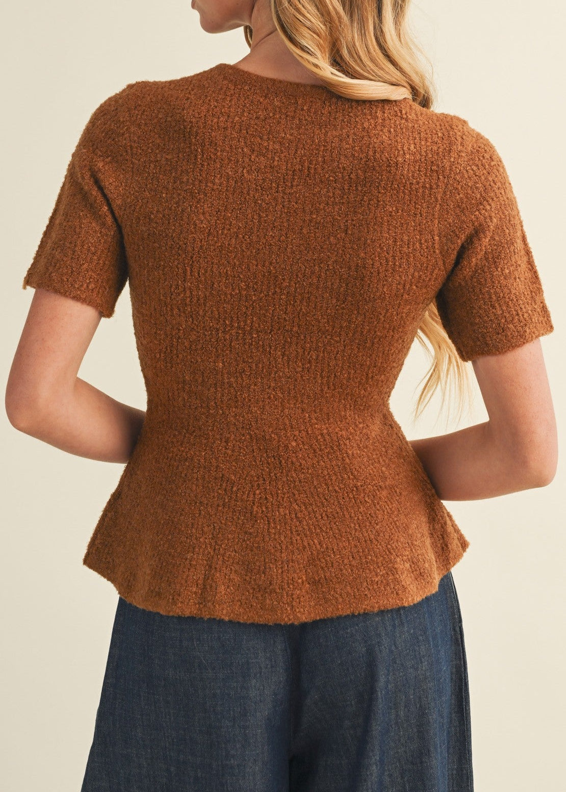 A woman shown from the back wearing a short-sleeve textured knit top in a warm rust brown color, paired with dark blue jeans, against a plain light beige background.