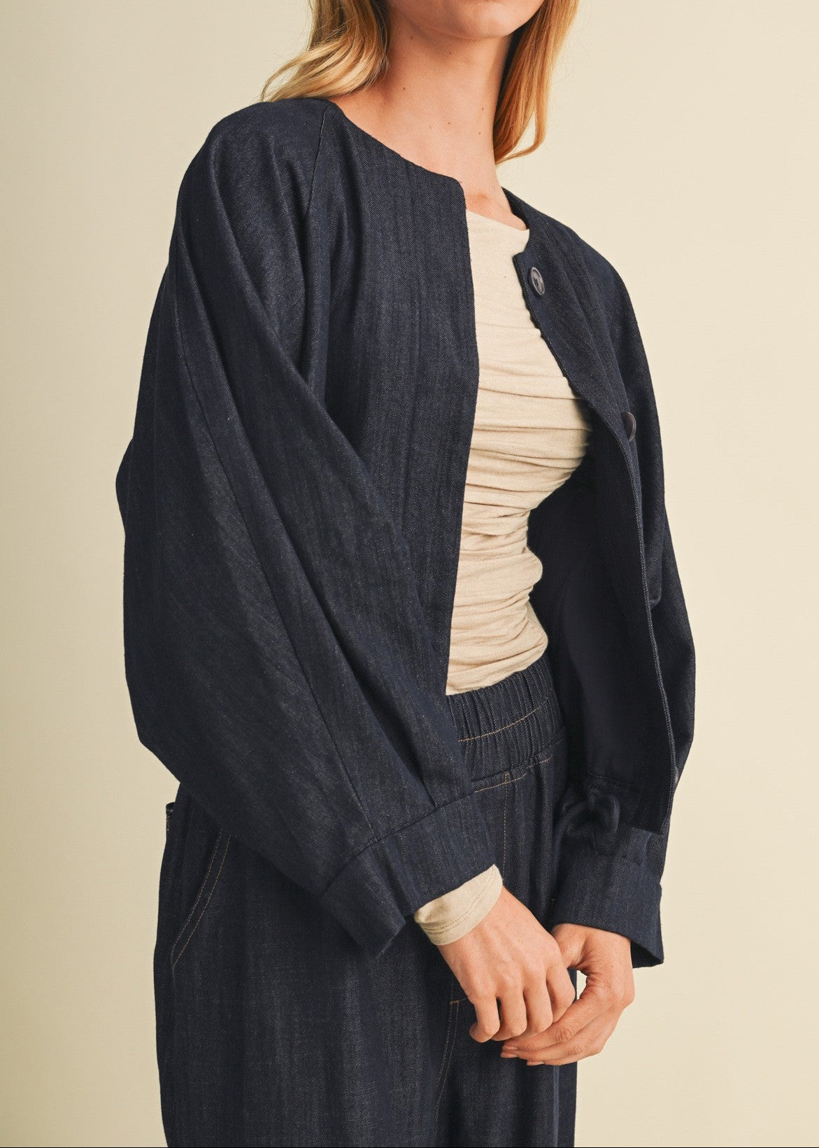 A woman modeling a dark navy blue linen-blend jacket with voluminous balloon sleeves, worn over a cream-colored ruched top and matching dark trousers, set against a solid warm beige studio background.