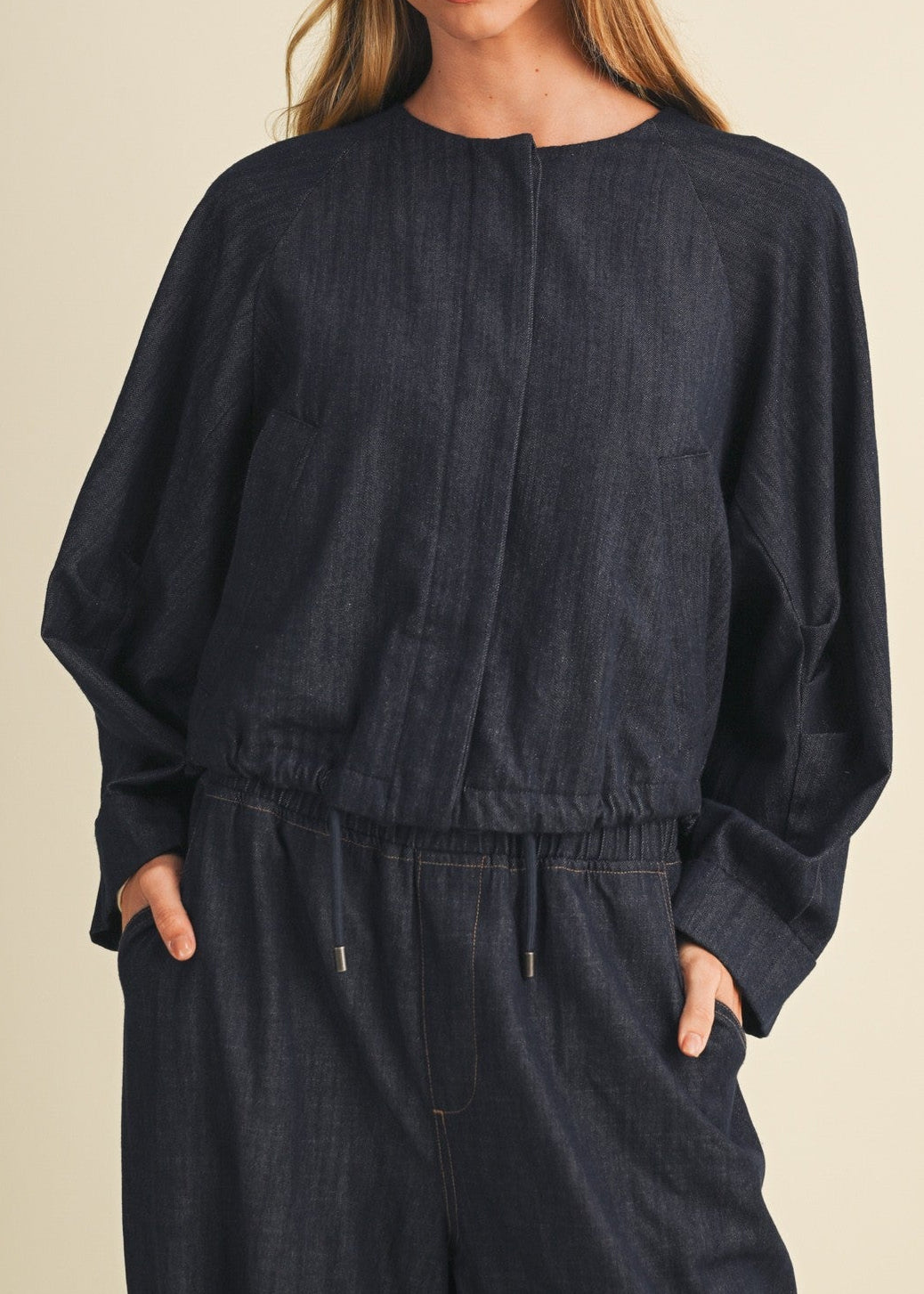 A woman modeling a dark navy blue linen jumpsuit with a drawstring waist and front pockets, set against a solid light beige background.