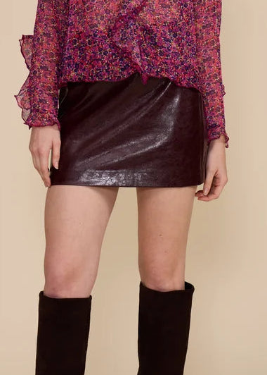 A model stands against a solid beige studio background wearing a long-sleeve ruffled blouse with a pink and purple floral pattern, a dark brown faux-leather mini skirt, and black knee-high boots.