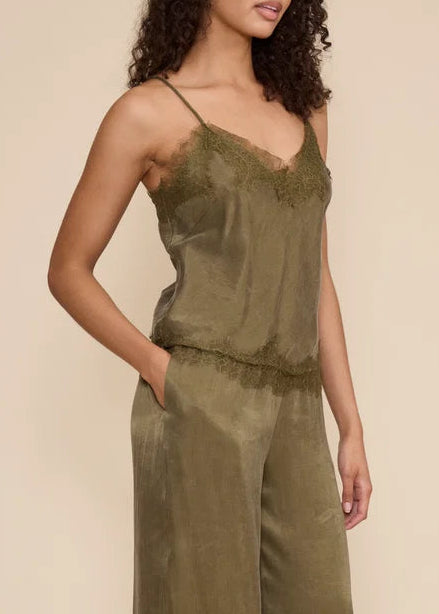 A woman modeling a sage green sleeveless jumpsuit with thin straps and wide-leg harem-style pants, standing against a plain beige studio background.