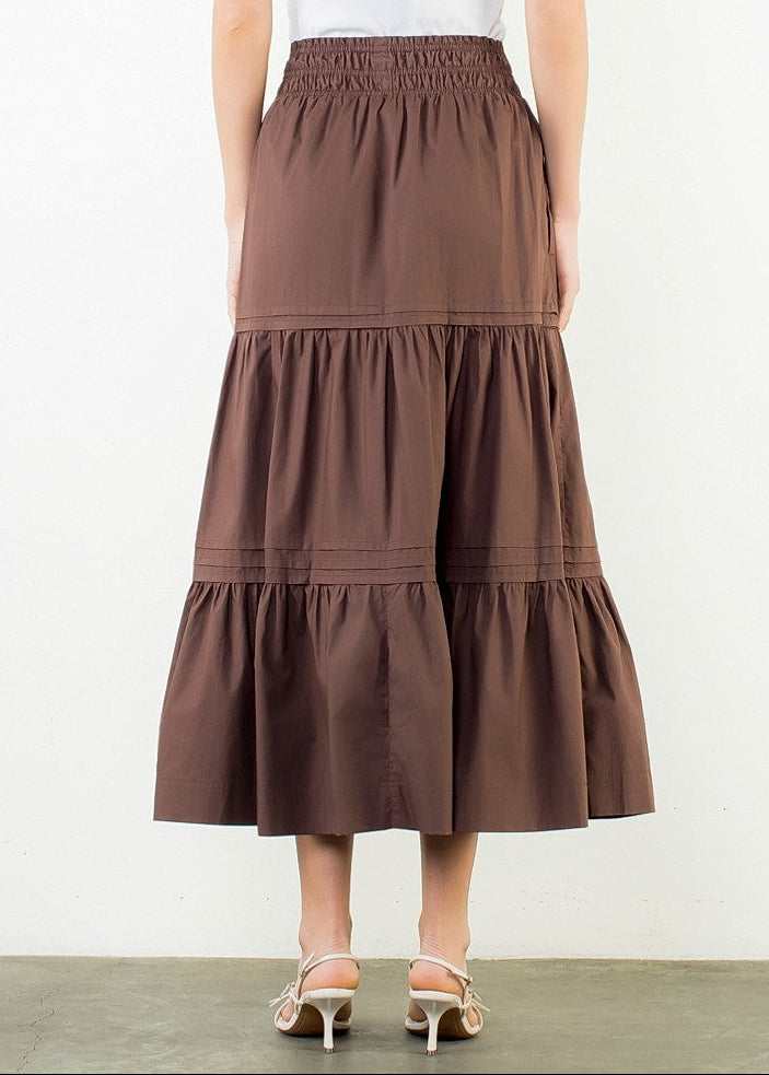 A woman shown from the back wearing a white short-sleeve top and a brown tiered midi skirt, standing against a plain white wall.