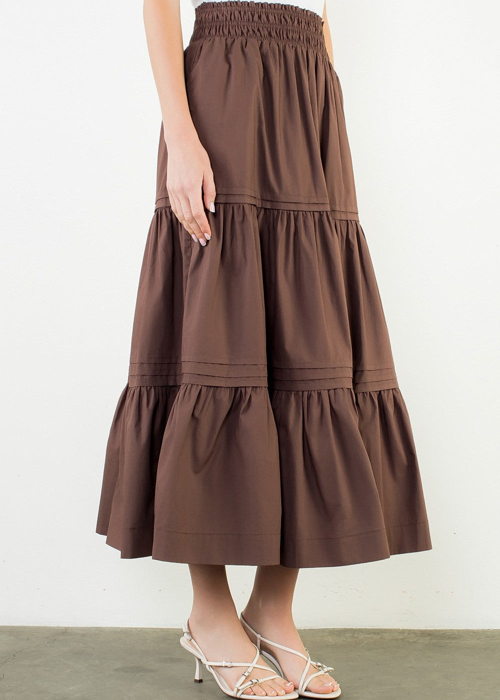 A brown tiered maxi skirt with an elastic waistband, shown on a model wearing a white top and silver heeled sandals against a plain white wall.