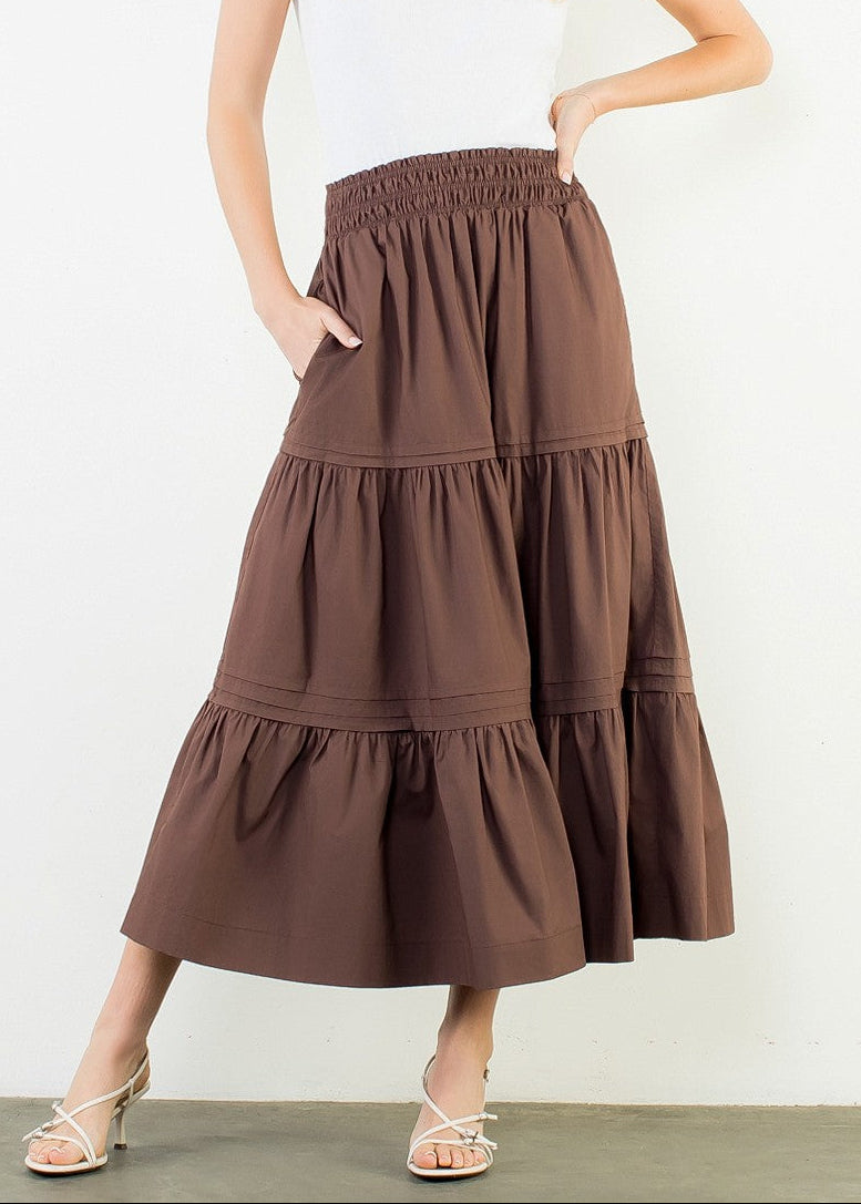 A woman modeling a brown tiered maxi skirt with side pockets, paired with a white t-shirt and beige strappy heels, standing against a plain white wall.