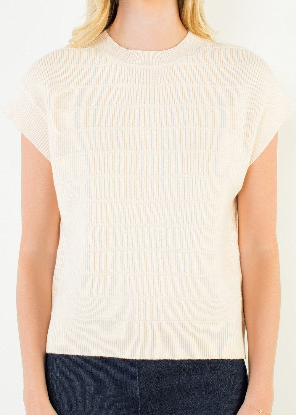 A woman wearing a cream-colored ribbed short-sleeve knit top and dark blue jeans, shown from the neck down against a plain white background.
